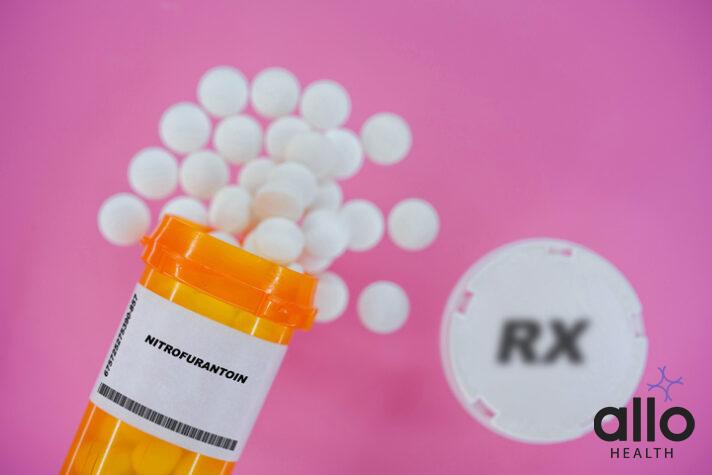 Nitrofurantoin Rx medicine pills in plactic vial with tablets. Pills spilling from yellow container on pink background.